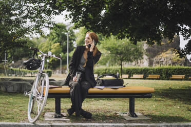 Happy businesswoman talking on mobile phone while sitting by bicycle at park - MASF34170