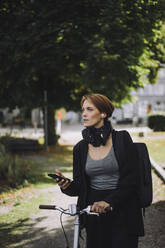 Female commuter with smart phone and bicycle walking in park - MASF34171