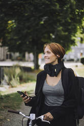 Glückliche Geschäftsfrau mit Mobiltelefon und Fahrrad beim Spaziergang im Park - MASF34172