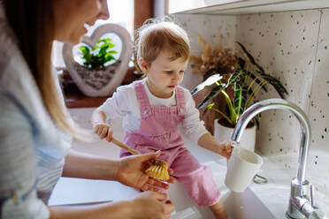 Kleines Mädchen sitzt auf der Küchentheke und hilft der Mutter, die Tasse in der Spüle in der Küche zu waschen. - HPIF03253