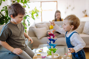 Kleine Geschwister spielen mit einer Montessori-Holzmurmelbahn im Wohnzimmer, ihre Mutter sitzt mit dem Laptop auf dem Sofa. - HPIF03261