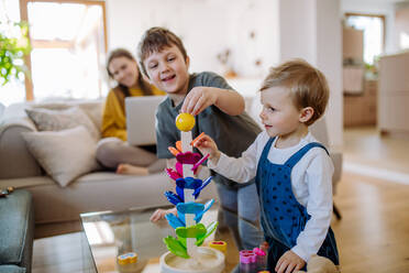 Kleine Geschwister spielen mit Holzspielzeug im Wohnzimmer, ihre Mutter sitzt auf einem Sofa mit Laptop. - HPIF03262