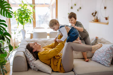 Eine junge, fröhliche Mutter spielt mit ihren kleinen Kindern und hat Spaß, wenn sie sie auf das Sofa hebt. - HPIF03270