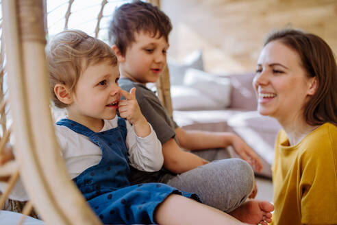 Eine junge, fröhliche Mutter, die mit ihren kleinen Kindern spielt und Spaß daran hat, sie auf einem Hängesessel im Wintergarten zu Hause zu schaukeln. - HPIF03276