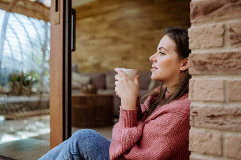 A smiling young woman with dreamy face drinking morning coffee and sitting at her home patio - HPIF03300