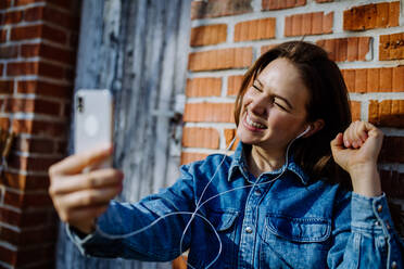 A happy young woman in denim jacket leaning the brick wall outdoors in summer and taking selfie - HPIF03307