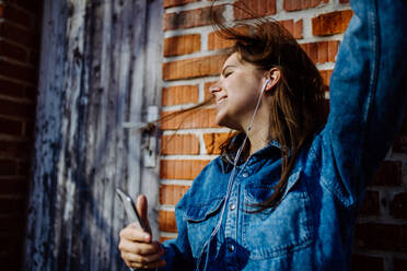 Eine glückliche junge Frau in Jeansjacke lehnt im Sommer draußen an der Backsteinmauer und hört Musik vom Smartphone. - HPIF03308