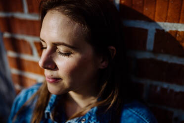 Portrait of happy young woman resting with closed eyes at home, in front of brick wall. - HPIF03309