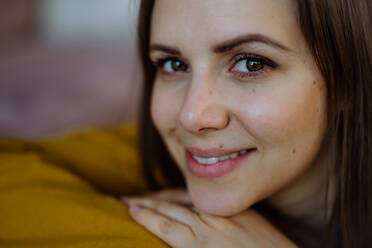 Ein Nahaufnahme-Porträt einer glücklichen jungen Frau, die lächelt und in die Kamera schaut, während sie sich zu Hause auf dem Sofa ausruht. - HPIF03313