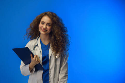 Eine Studioaufnahme einer jungen Ärztin, die vor einem blauen Hintergrund in die Kamera schaut. - HPIF03419
