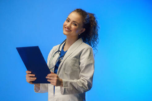Eine Studioaufnahme einer jungen Ärztin, die vor einem blauen Hintergrund in die Kamera schaut. - HPIF03420