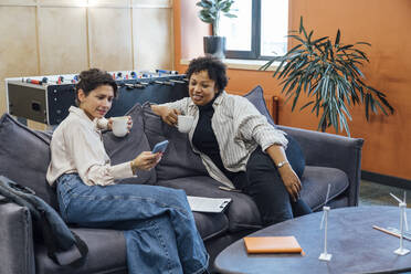 Lächelnde Geschäftsfrau mit einem Kollegen, der ein Smartphone auf dem Sofa im Büro benutzt - VPIF07714