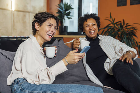 Glückliche Geschäftsfrauen mit Kaffeetassen auf dem Sofa im Büro sitzend - VPIF07717