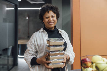 Happy young woman with take away food boxes standing in office - VPIF07748