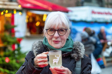 Lächelnde Seniorin mit einer Tasse Glühwein auf dem Weihnachtsmarkt - FRF01004