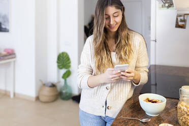 Glückliche junge Frau benutzt ihr Smartphone in der Küche zu Hause - JPTF01165