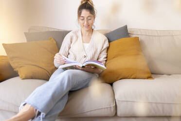 Smiling young woman reading book on sofa at home - JPTF01173