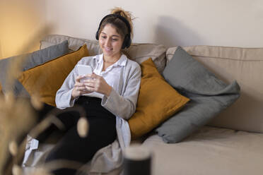 Happy young woman wearing wireless headphones using smart phone on sofa at home - JPTF01176