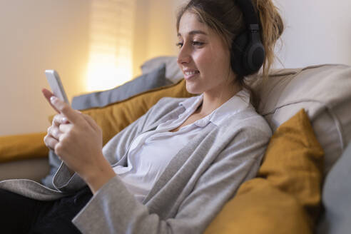 Lächelnde Frau mit drahtlosen Kopfhörern und Smartphone auf dem Sofa zu Hause - JPTF01177