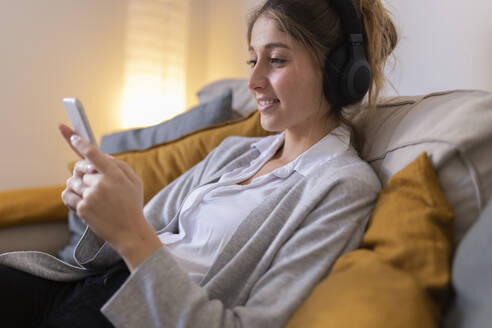 Smiling woman wearing wireless headphones using smart phone on sofa at home - JPTF01177