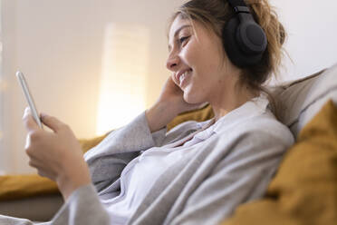 Happy woman wearing wireless headphones using smart phone at home - JPTF01178