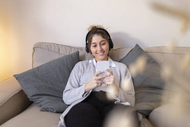 Happy woman wearing wireless headphones using smart phone on sofa at home - JPTF01179