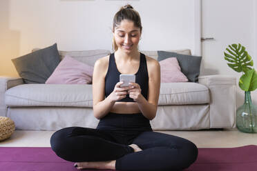 Lächelnde Frau benutzt ihr Smartphone auf einer Matte zu Hause - JPTF01196