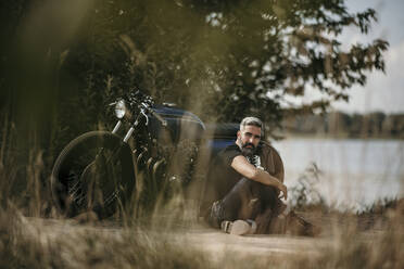 Nachdenklicher reifer Mann sitzt vor einem Motorrad - MJRF00800