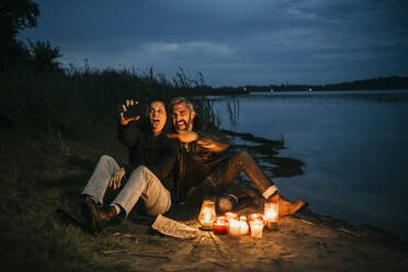 Glückliche Frau, die ein Selfie mit einem Mann macht, der an einem Seeufer bei Kerzenschein sitzt - MJRF00827