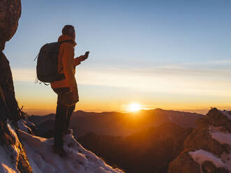 Pensive hiker using smart phone on mountain peak during sunrise at