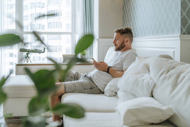 Junger Mann mit Bart benutzt ein Smartphone auf dem Sofa zu Hause - MDOF00368
