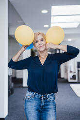 Lächelnde reife Geschäftsfrau hält Luftballons am Arbeitsplatz - JOSEF15218