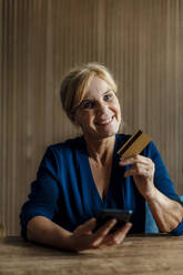 Happy businesswoman holding mobile phone and credit card in office - JOSEF15238