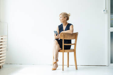 Geschäftsfrau mit Smartphone sitzt auf einem Stuhl vor einer weißen Wand - JOSEF15242