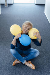 Geschäftsfrau mit geschlossenen Augen, die Luftballons auf dem Boden sitzend im Büro hält - JOSEF15252