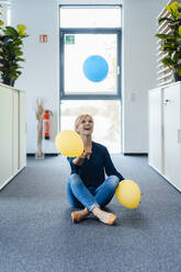 Glückliche Geschäftsfrau, die mit Luftballons spielt, die auf dem Boden am Arbeitsplatz sitzen - JOSEF15255