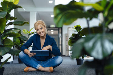 Happy businesswoman with tablet PC sitting on ground in office - JOSEF15258