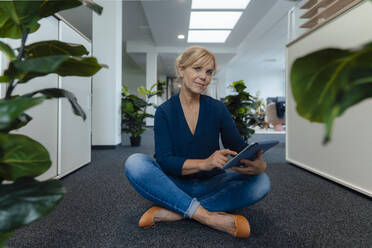 Mature businesswoman holding tablet PC sitting on ground in office - JOSEF15262