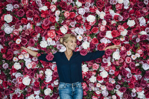 Happy businesswoman with arms outstretched leaning on flower hedge - JOSEF15265