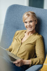 Glückliche reife Geschäftsfrau mit Laptop auf einem Sessel - JOSEF15312