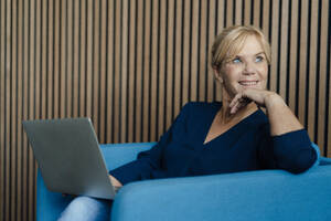 Glückliche Geschäftsfrau mit Laptop auf einem Sessel - JOSEF15362