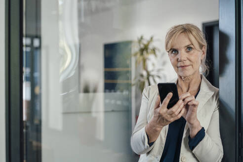 Reife Geschäftsfrau mit Mobiltelefon im Büro durch Glas gesehen - JOSEF15370