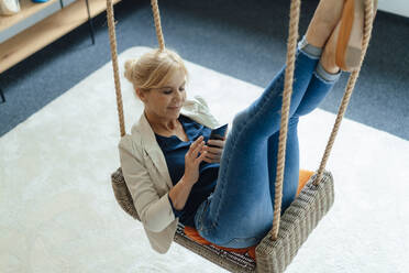 Businesswoman using mobile phone on swing at workplace - JOSEF15377