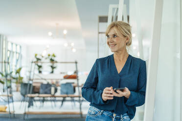 Happy businesswoman with mobile phone leaning on wall in office - JOSEF15382