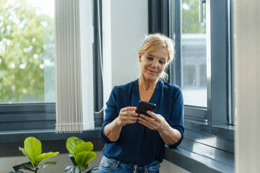 Reife Geschäftsfrau benutzt Smartphone am Fenster im Büro - JOSEF15391