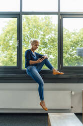 Businesswoman using smart phone sitting on window sill at office - JOSEF15395