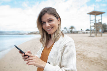 Lächelnde schöne junge Frau mit Smartphone am Strand - JOSEF15436