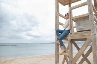 Smiling young woman sitting on wooden lifeguard hut at beach - JOSEF15438