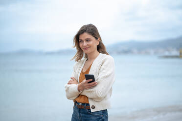 Lächelnde schöne junge Frau mit verschränkten Armen hält Smartphone am Strand - JOSEF15445