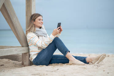 Lächelnde schöne junge Frau, die ein Smartphone benutzt und sich am Strand an ein Holz lehnt - JOSEF15447